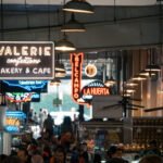 Indoor view of a bustling marketplace with neon signs for "Valerie Bakery" and other vendors.