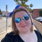 Person with American flag headband, sunglasses, and star earrings smiling outside on a sunny day.